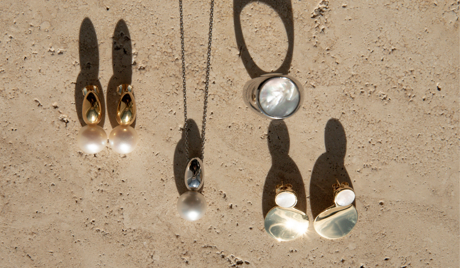 The Ebb collection sitting on a travertine surface, featuring Mother of Pearl, South Sea pearl pieces set in sterling silver and gold vermeil with high polish finishes.