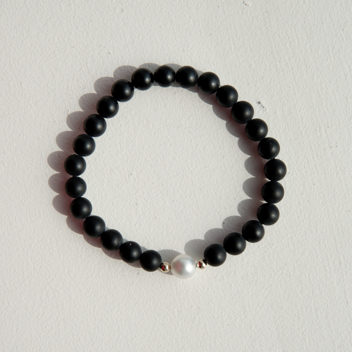 A black onyx bracelet with a near round Australian South Sea pearl centerpiece, threaded with elastic and placed on a textured stone surface.