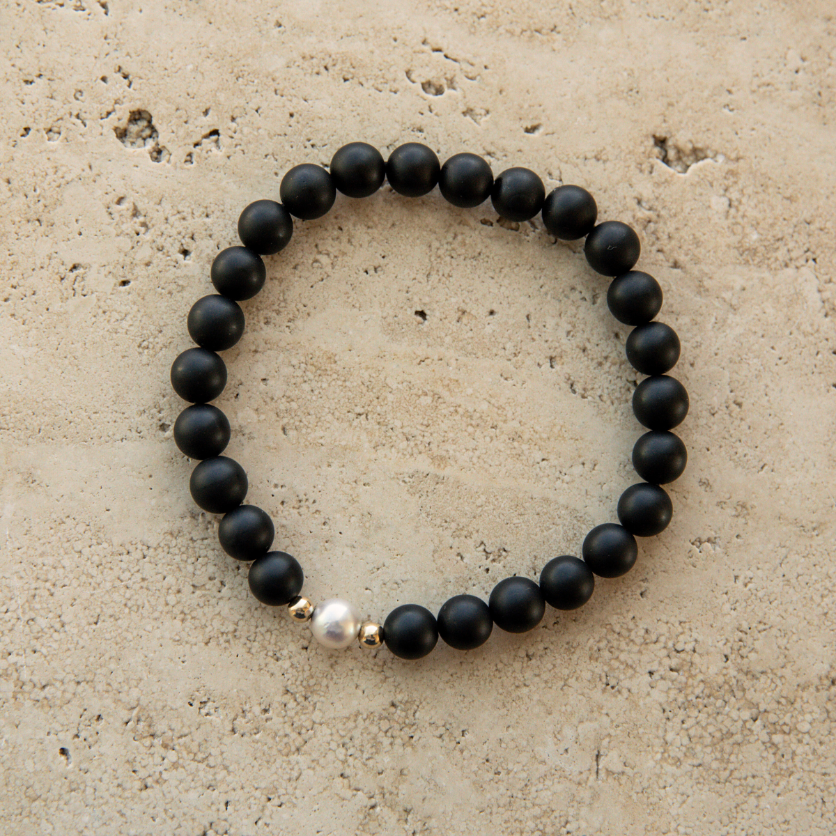 Black onyx beaded bracelet with a pearl and gold accent on a beige stone surface