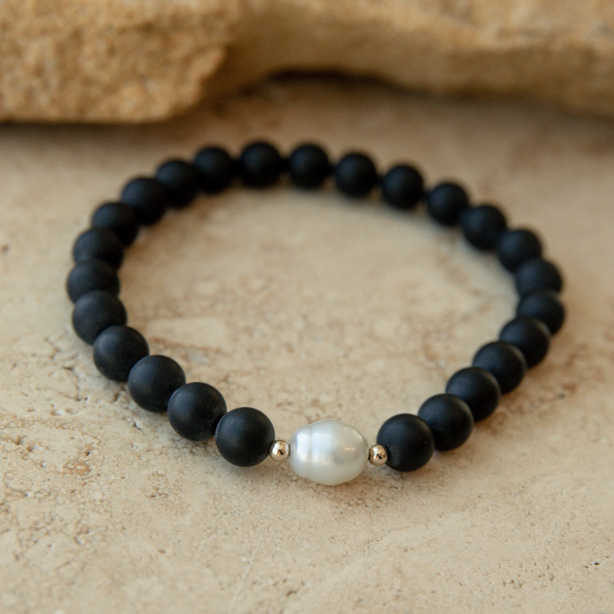 Black onyx beaded bracelet with a white Australian South Sea pearl on a beige stone surface.