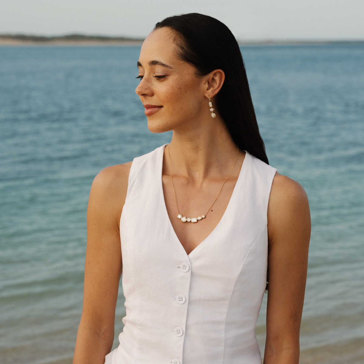 Woman in a white sleeveless top standing by the water wearing a mother of pearl necklace and earrings