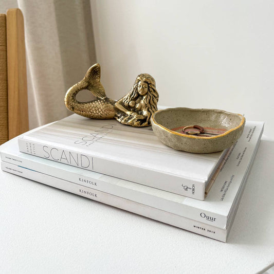 Decorative setup with a gold mermaid figurine, ceramic bowl, and books on a light surface.