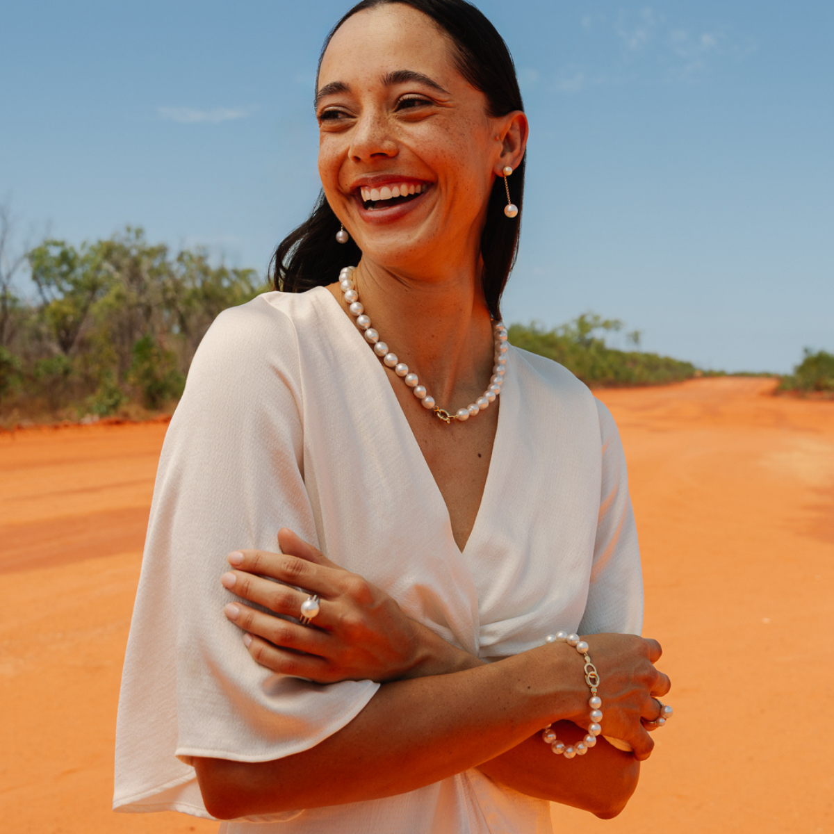 Woman in a white dress with jewelry standing on a red dirt road with a clear blue sky.