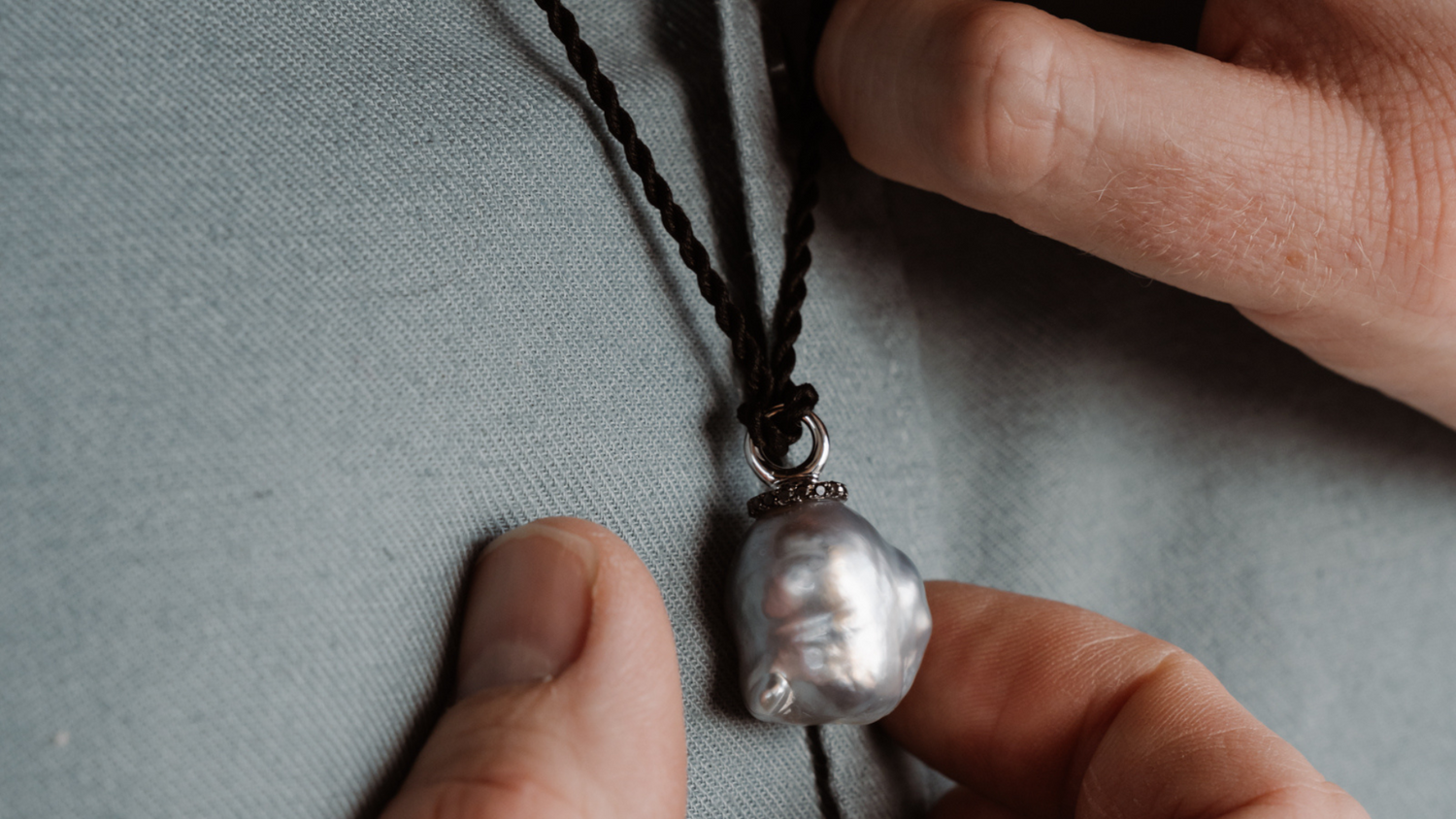 A man holds his stunning pendant that features an Australian South Sea Baroque pearl set alongside black diamonds, and hung on a black silk cord necklace.