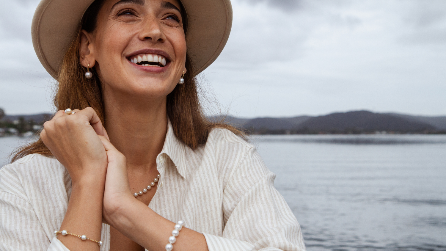 A woman broadly smiles wearing her Australian Saltwater pearl jewellery with the Broken Bay Pearl Farm in the background of the photo.