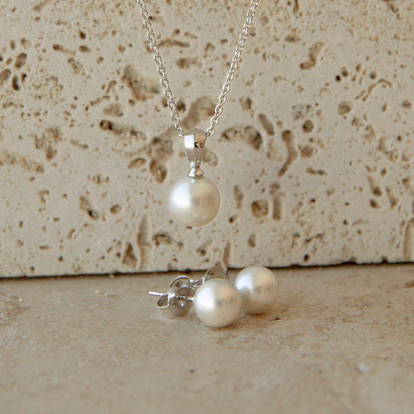 A silver necklace with a single round white Akoya pearl pendant and matching Akoya pearl stud earrings, displayed on a textured beige stone surface.