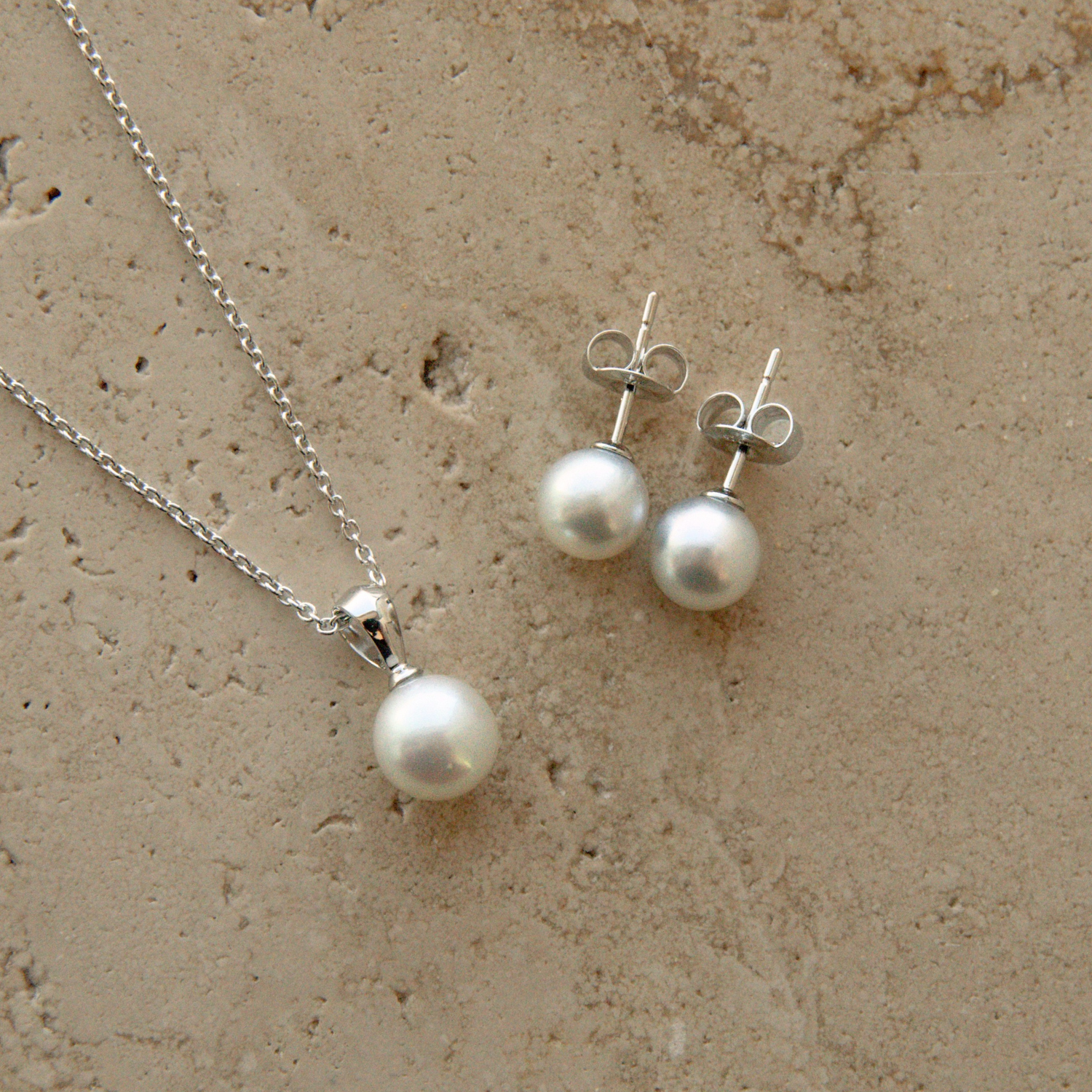 A silver necklace and matching stud earrings with white Akoya pearls, displayed on a textured beige stone surface.