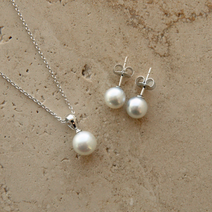 A silver necklace and matching stud earrings with white Akoya pearls, displayed on a textured beige stone surface.