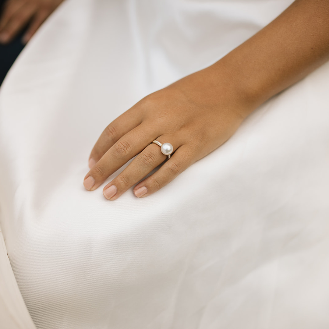 A bride rests her hand on her stunning white gown, and on her wedding finger she wears a gorgeous pearl ring with a band of white sparkling diamonds set alongside the pearl.