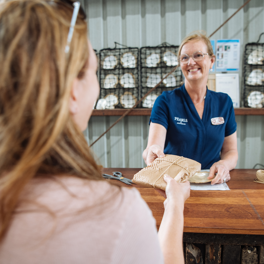 Our friendly pearl expert stands at the point of sale of our Shellar Door, handing over a beautiful Mother of Pearl shell purchased by a visiting customer. She smiles broadly and the customer is excited to take her memento from her time at Broken bay pearl farm home with her.