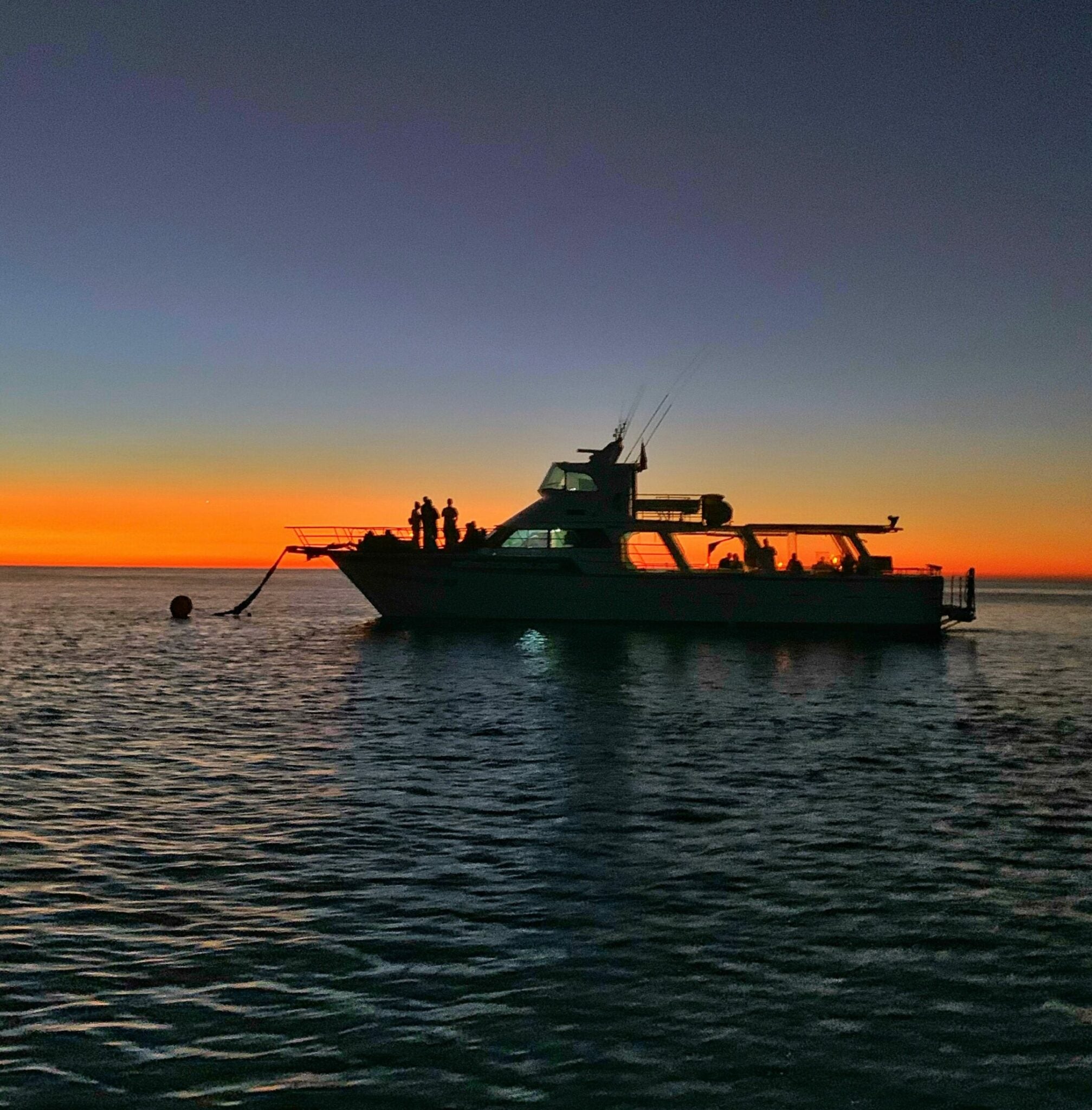 A boat sits on the tranquil Kimberley waters at sunset, with the sky illuminated by a warm orange glow.