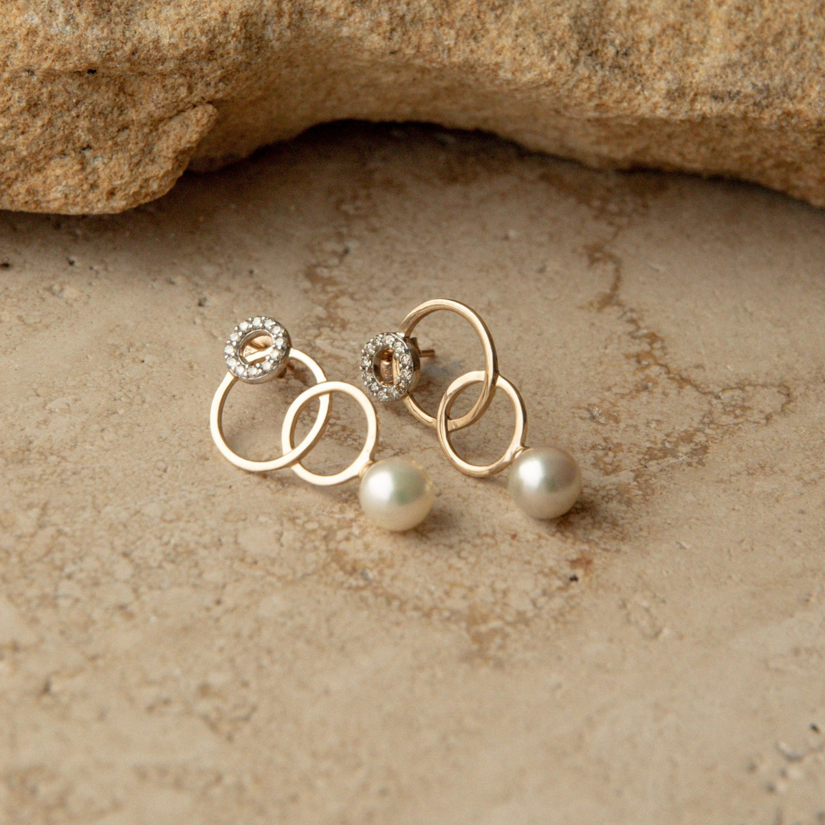 A pair of gold earrings with interlocking circular rings, one adorned with a small white diamond clusters and the other with a single Australian Akoya pearl drop, displayed on a textured beige stone surface.