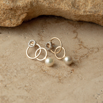 A pair of gold earrings with interlocking circular rings, one adorned with a small white diamond clusters and the other with a single Australian Akoya pearl drop, displayed on a textured beige stone surface.