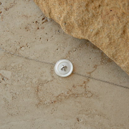A delicate silver chain bracelet featuring a round white Mother of Pearl button motif, displayed on a textured beige stone surface next to a larger tan rock.