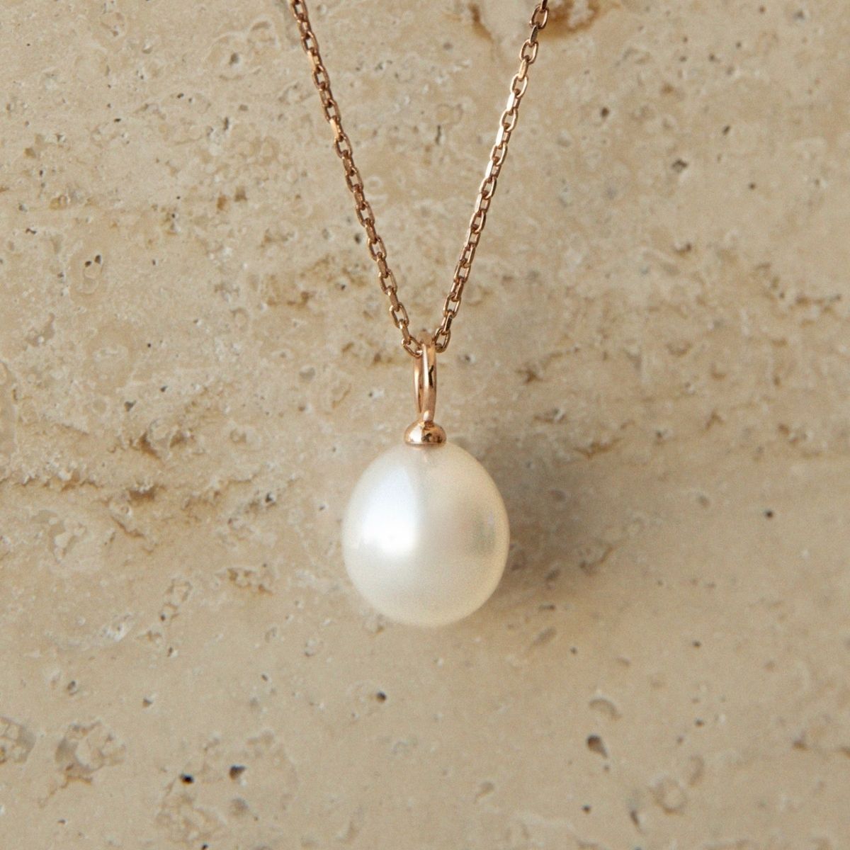 A delicate rose gold chain necklace featuring a single lustrous white Australian South Sea pearl charm, displayed against a textured beige stone background.