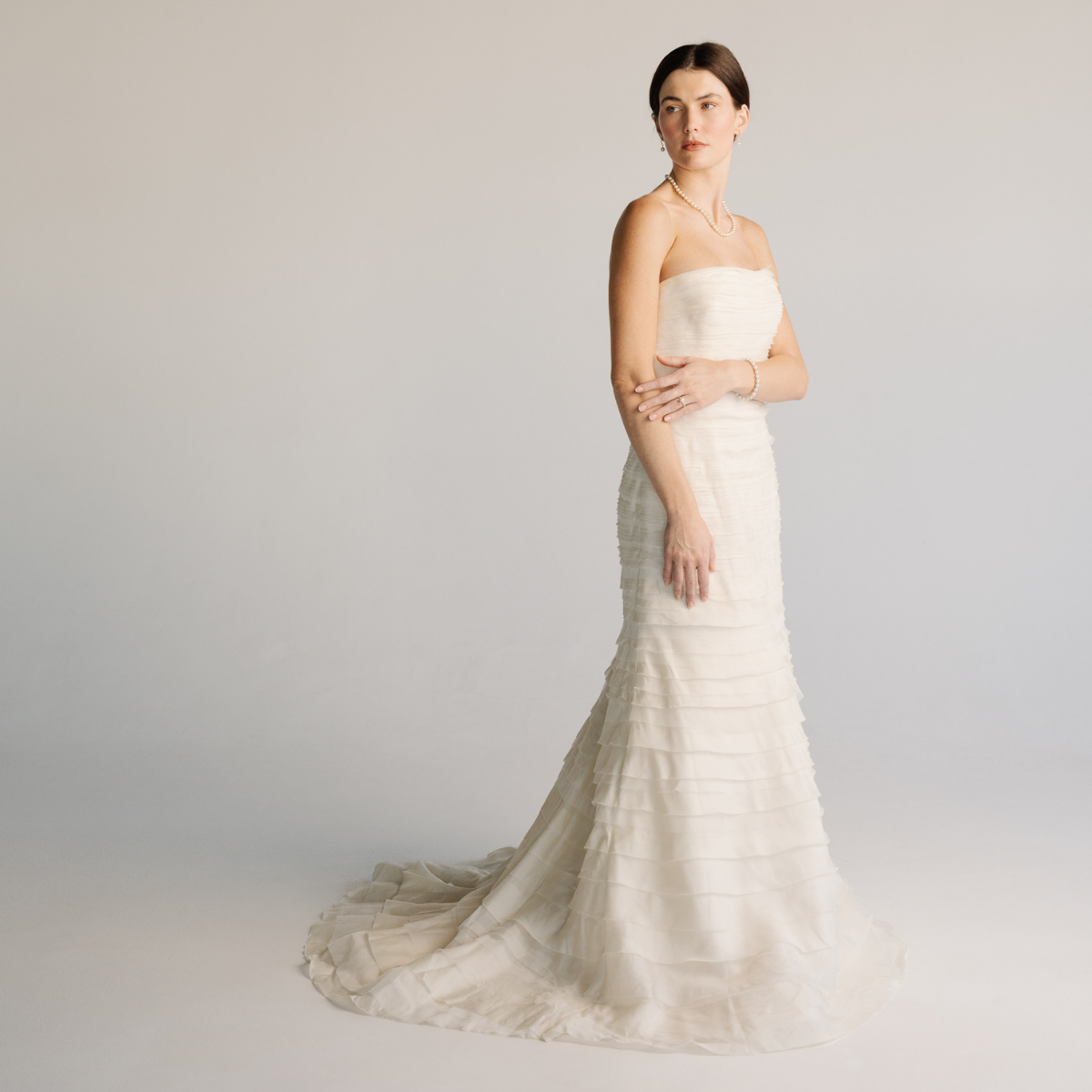  dress and a matching bridal jewellery set of a pearl strand necklace, bracelet, earrings, and a ring while standing against a plain background.