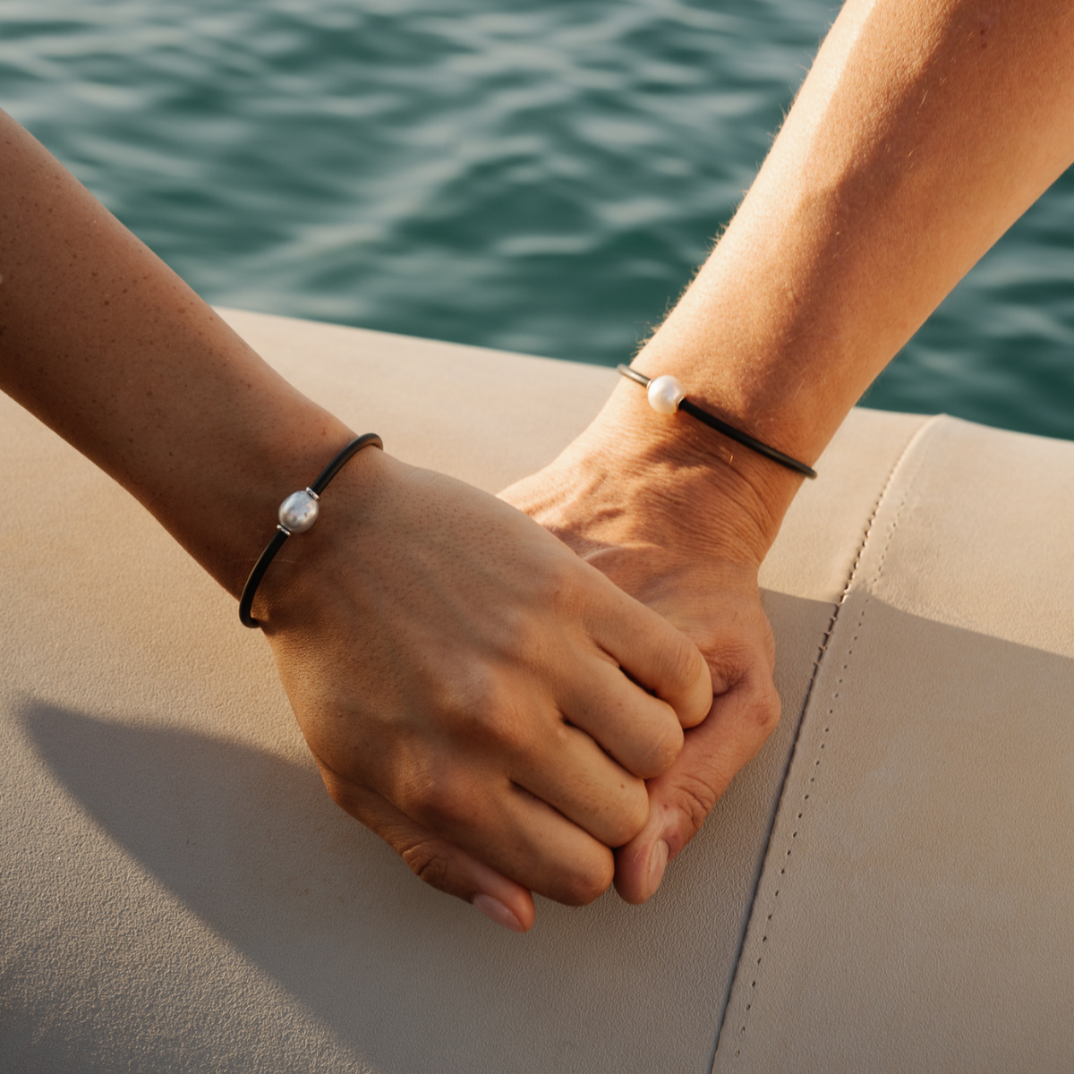 Two hands holding each other, each wearing a classic pearl neoprene bracelet with a blurred water background of our Cygnet Bay Pearl Farm.