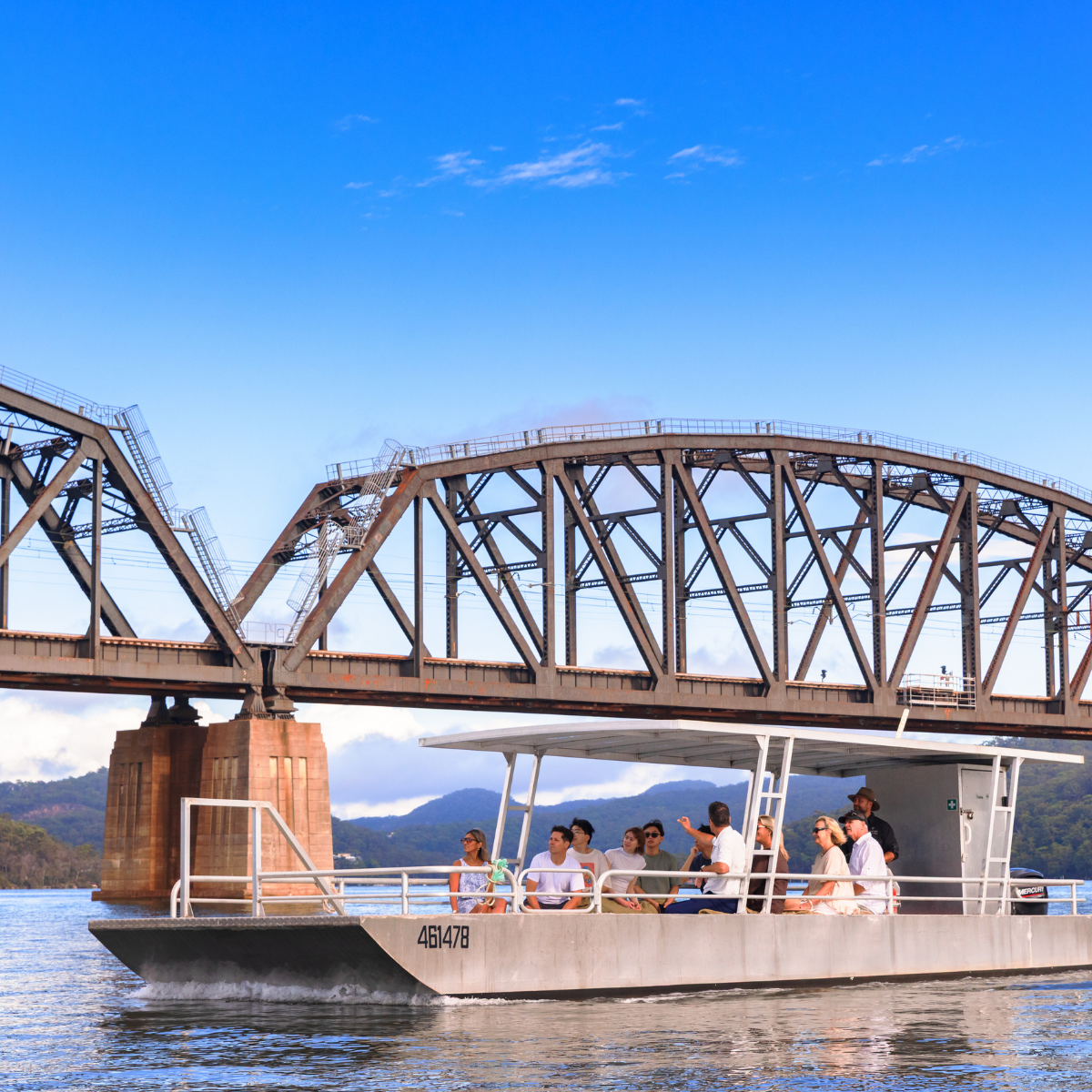 Boat with passengers from the Shellar Door at Broken Bay Pearl Farm on the lower Hawkesbury River under a bridge with ranges in the background.