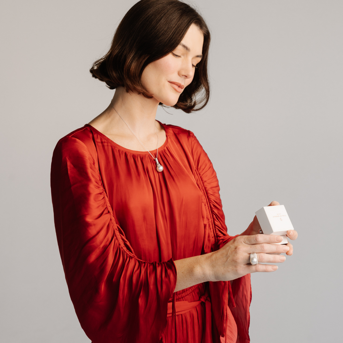 Woman in a red dress holding a white Pearls of Australia box against a gray background. She wears a stunning pair of Stella pearl earrings, ring and pendant on a chain.