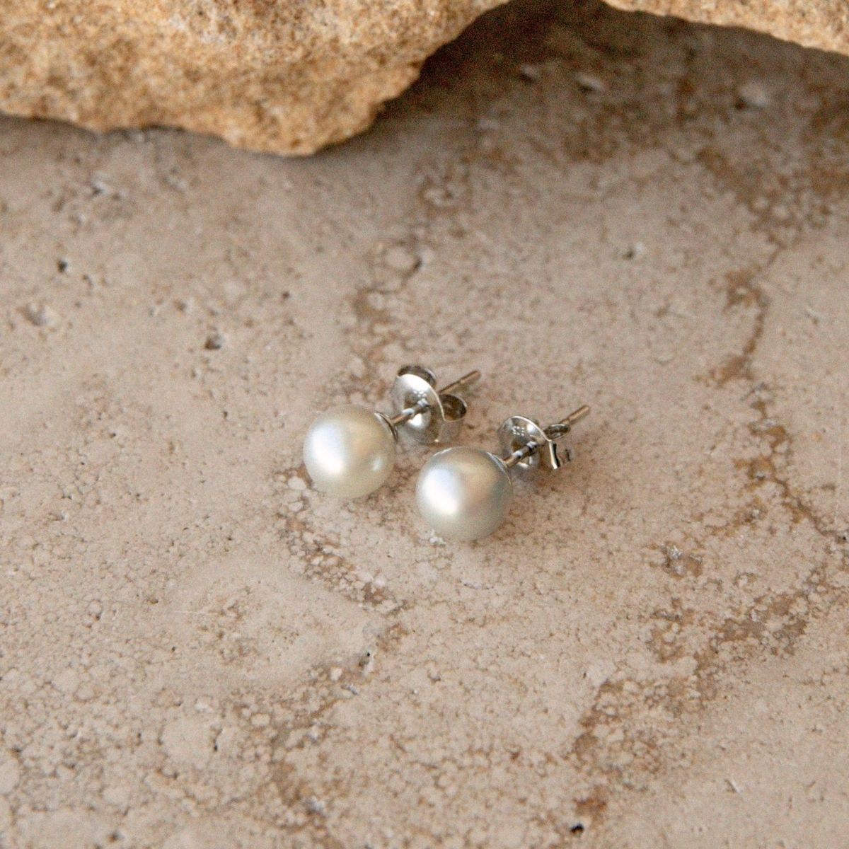 A pair of silver Akoya pearl stud earrings, resting on a textured sandy surface with a rock in the background.