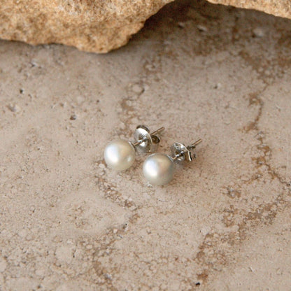 A pair of silver Akoya pearl stud earrings, resting on a textured sandy surface with a rock in the background.