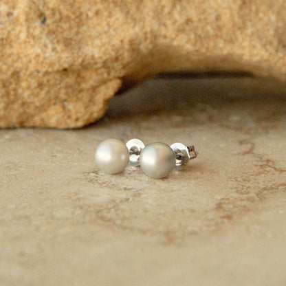 A pair of silver stud earrings with AA grade, round, lustrous Australian Akoya pearls, resting on a textured beige stone surface with a larger rock in the background.