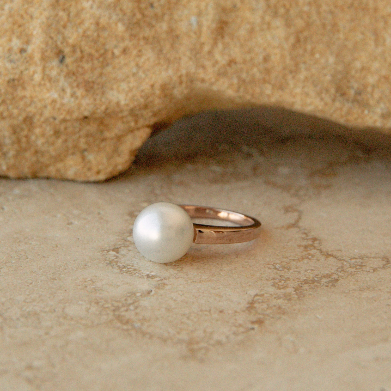 A single ring featuring a round white Australian South Sea pearl set in a polished rose gold band, resting on a textured beige stone surface with a larger tan rock in the background.