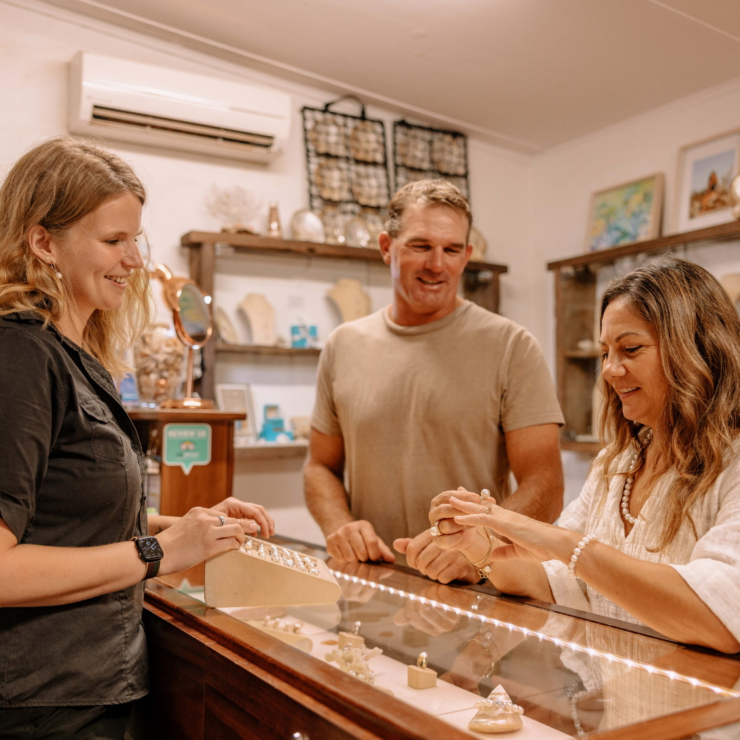 A couple stand at the front cabinet of the Cygnet Bay Gallery, trying on stunning pearl jewellery pieces set in gold. Each piece features a beautiful Australian Saltwater pearl grown at one of our pearl farms.