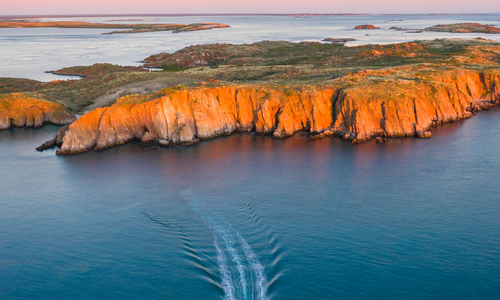 A beautiful aerial photo of a group of guests on a Sea Safari tour at sunset, with the islands glowing orange in the last light of the day. The sky has a pastel gradient, contrasting with the deep blue waters of the Kimberley.