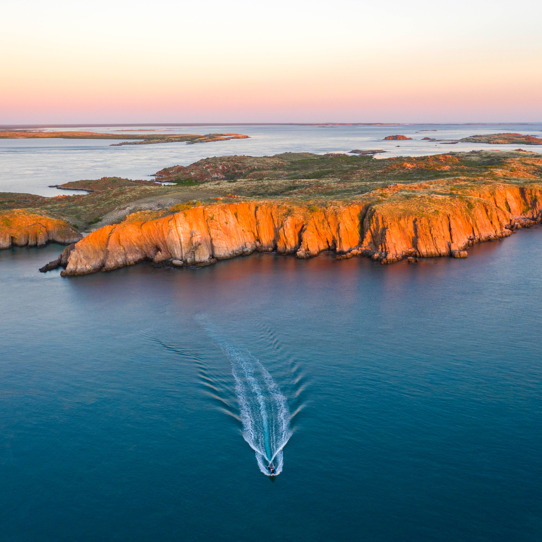 A beautiful aerial photo of a group of guests on a Sea Safari tour at sunset, with the islands glowing orange in the last light of the day. The sky has a pastel gradient, contrasting with the deep blue waters of the Kimberley.