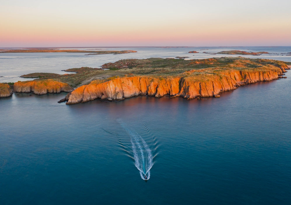 A boat travels along the calm, turquoise waters of the Kimberley at sunset. Behind them is a beautiful island, glowing in the warm orange glow of the sunset. The sky has a beautiful pink gradient.