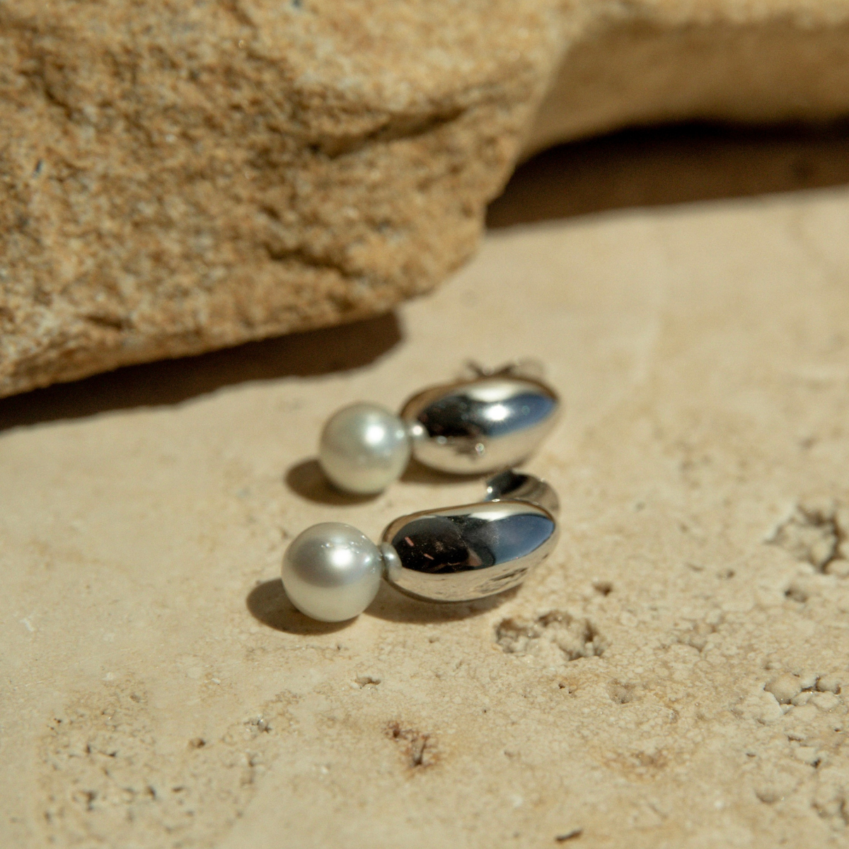 Silver earrings featuring a stunning Australian grown Akoya pearl resting on a sandy surface with a rock in the background