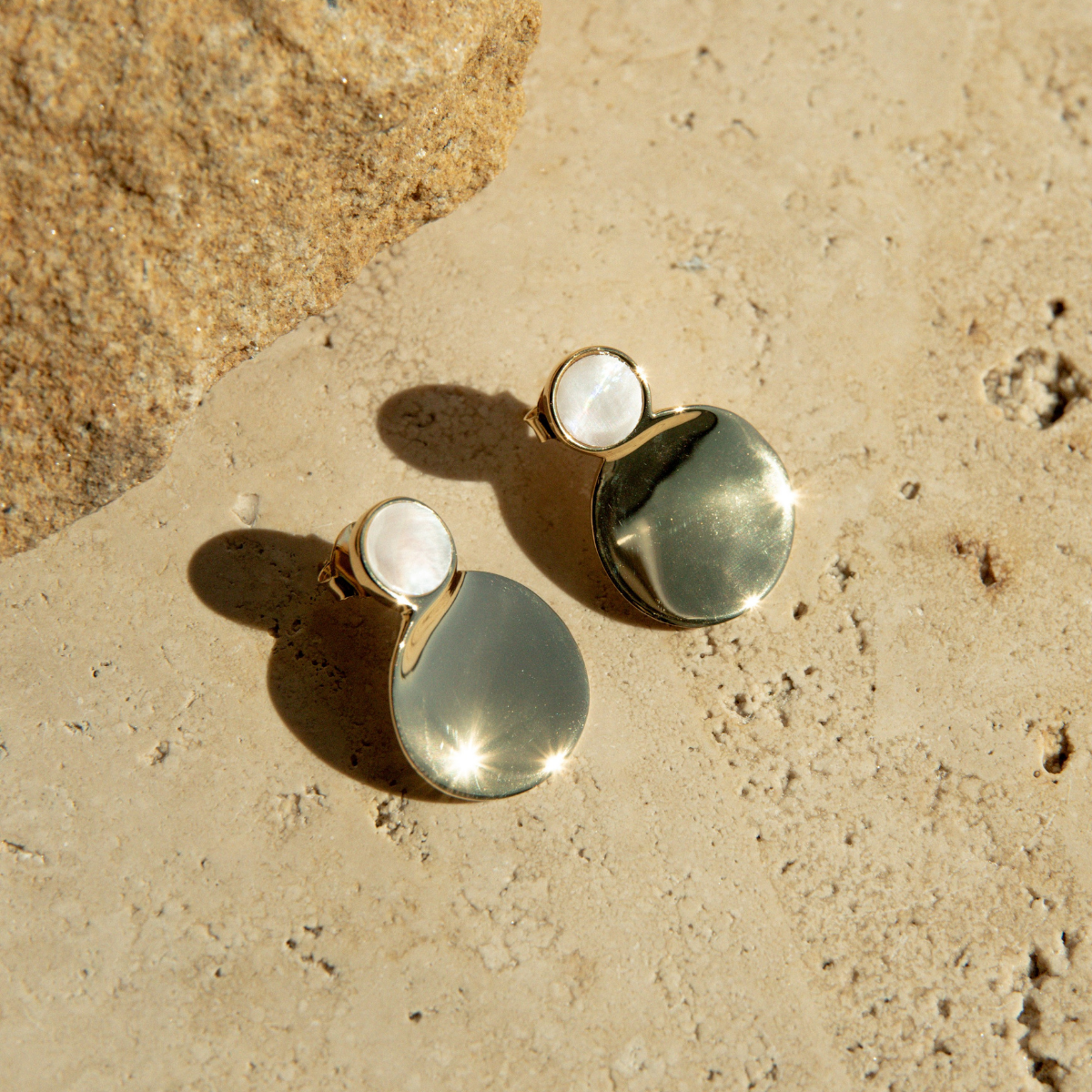 Pair of gold plated and mother of pearl earrings on a textured beige surface.
