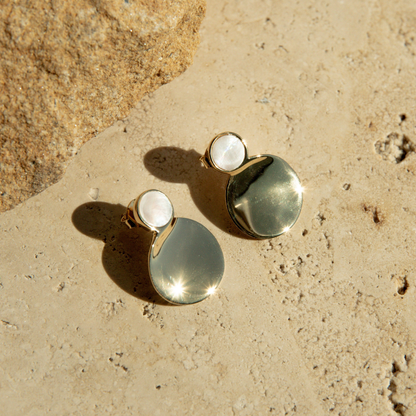 Pair of gold plated and mother of pearl earrings on a textured beige surface.