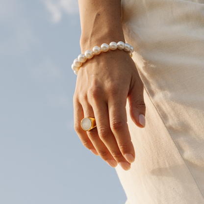 Hand wearing a pearl bracelet and mother of pearl signet ring against a blurred natural background