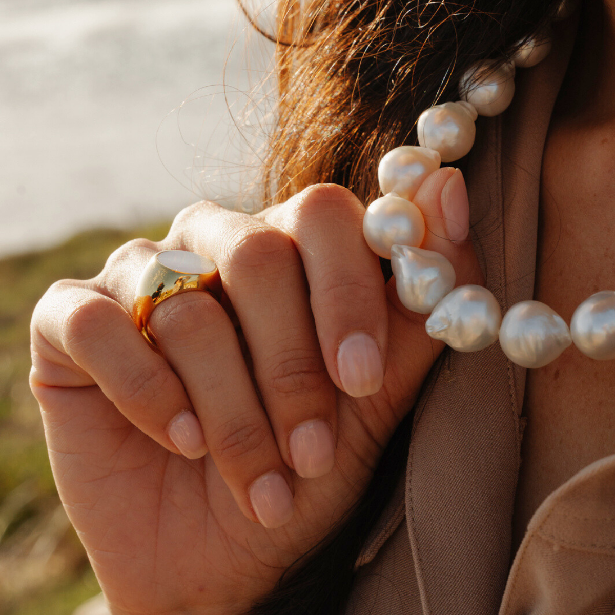 Close-up of a person wearing a pearl necklace and a mother of pearl signet ring with a blurred natural background