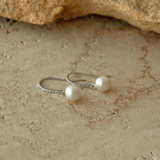 A pair of white gold and diamond hook earrings with round white Australian Akoya pearls, resting on a textured sandy surface with a rock in the background.