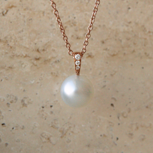 A rose gold necklace with a single Australian South Sea pearl pendant with three white diamonds on the bail, displayed against a textured beige stone background.