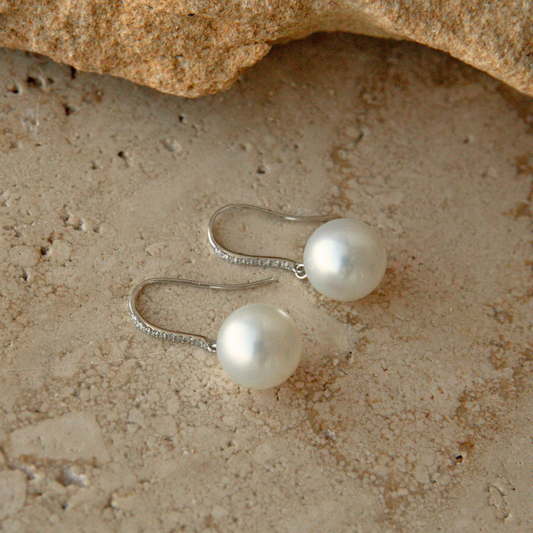 A pair of white gold and diamond hook earrings with round white Australian South Sea pearls, resting on a textured beige stone surface with a rock at the top.