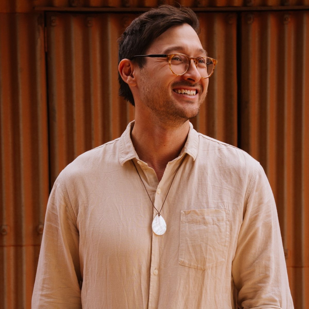 A man wearing a light beige linen button-down shirt, a white mother of pearl pendant necklace, and tortoiseshell glasses, standing in front of a warm brown corrugated metal wall.