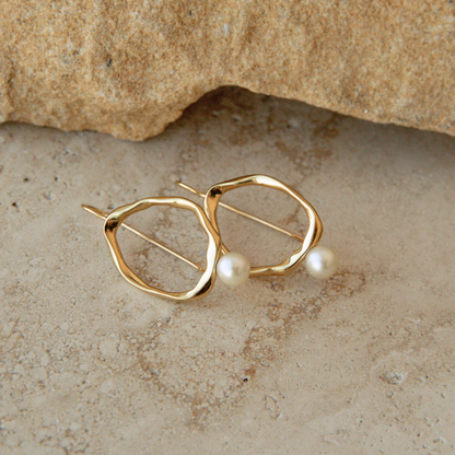 A pair of gold vermeil abstract hoop earrings with small white Akoya pearls, displayed on a textured beige stone surface.