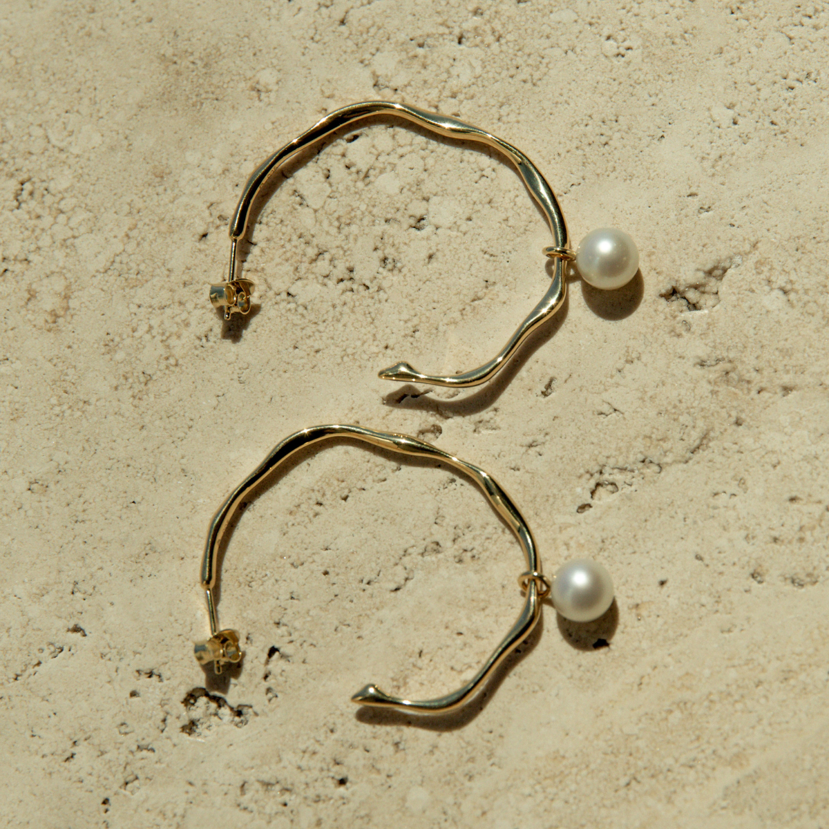 Gold hoop earrings with pearl drops on a beige stone surface