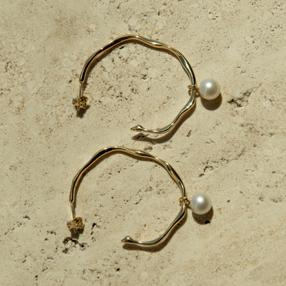 Gold hoop earrings with pearl drops on a beige stone surface