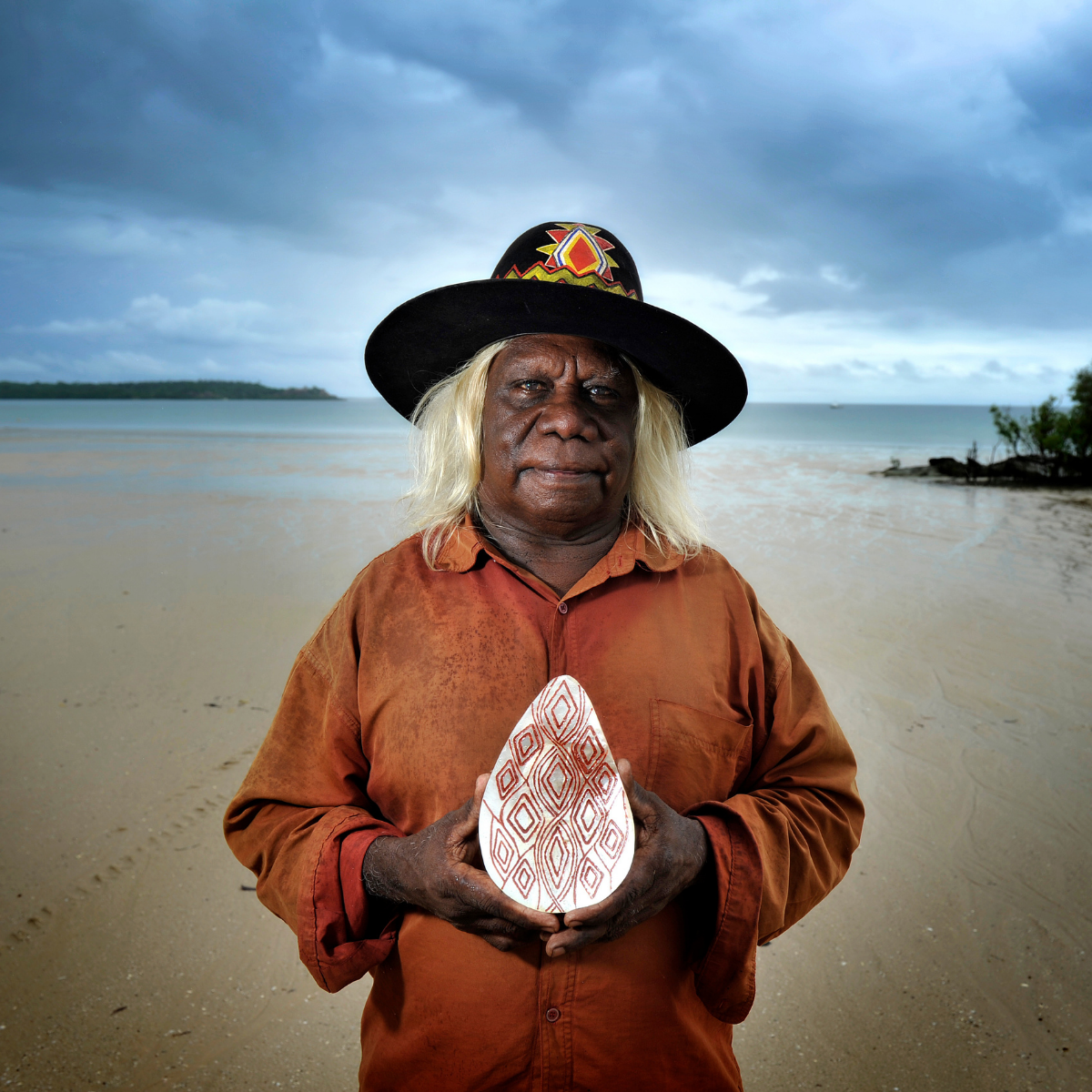 Bruce Wiggan, Master Pearl Shell Carver, stands on a sandy beach holding a carved Riji shell with red geometric patterns. He is wearing a wide-brimmed black hat with colorful beadwork and a rust-orange shirt, with the ocean and cloudy sky in the background.