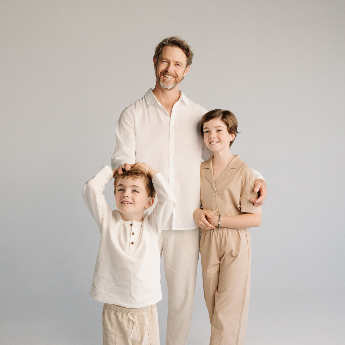 A family portrait featuring James Brown and his son and daughter wearing matching linen clothing sets in off-white and beige, standing against a solid light gray background.