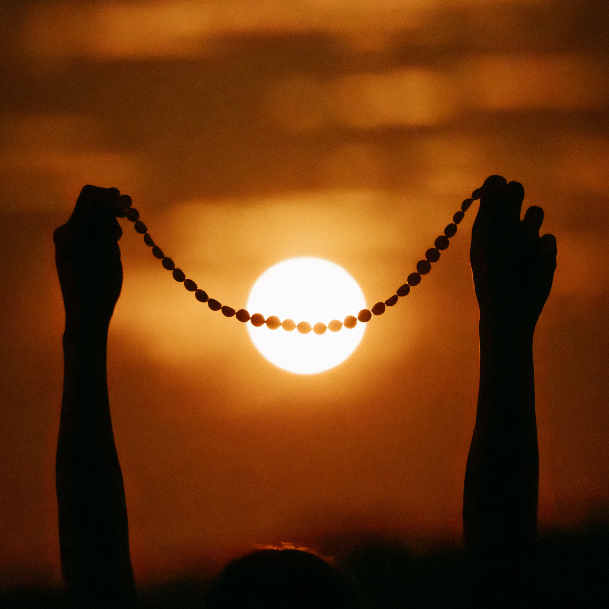 Silhouetted hands holding a string of Mabe pearls in front of a glowing sunset at Cygnet Bay Pearl Farm, with the sun low on the horizon and warm orange light filling the frame.
