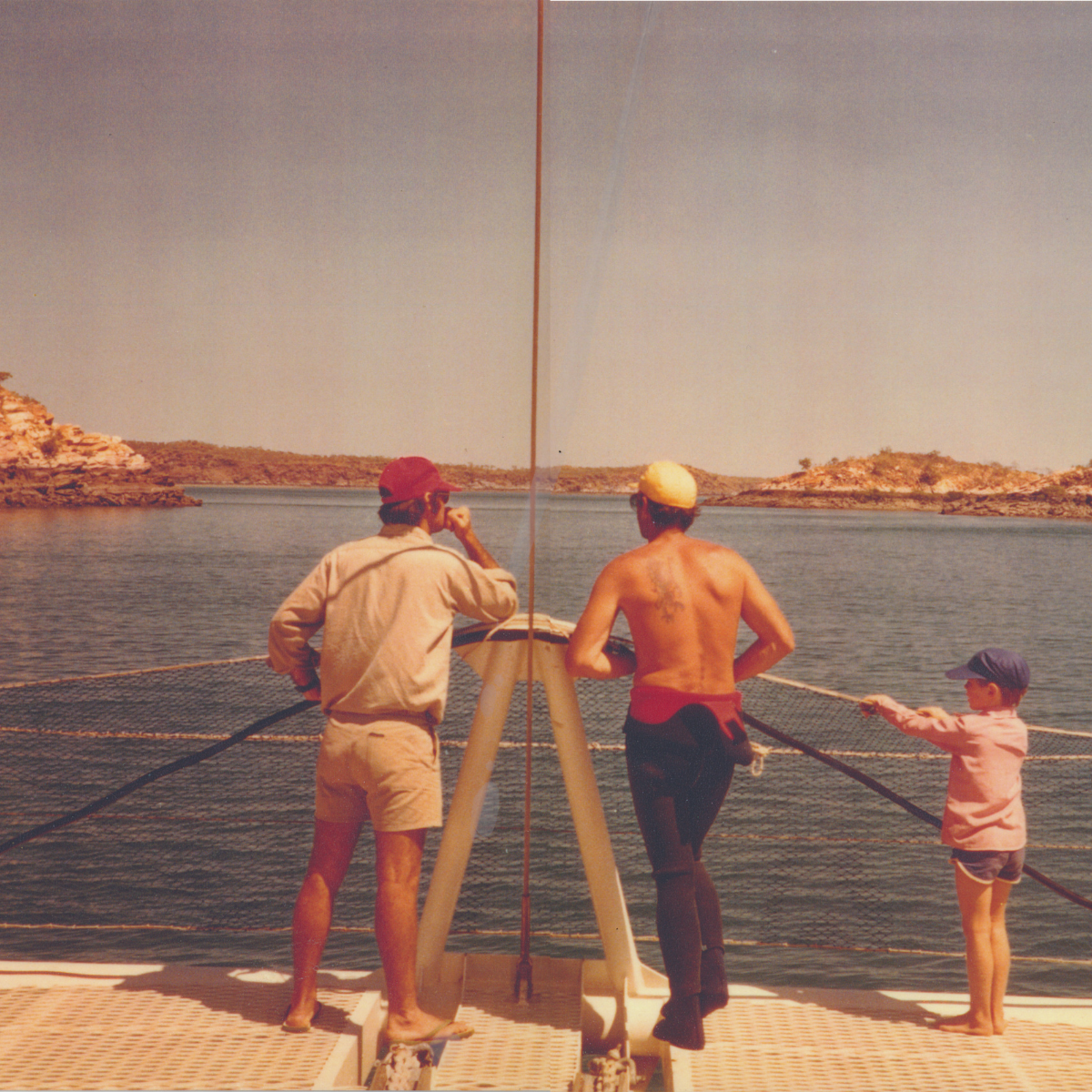 A vintage photograph showing Bruce Brown, a diver and young James Brown on the deck of a boat looking out over a body of water toward rocky shoreline islands. 