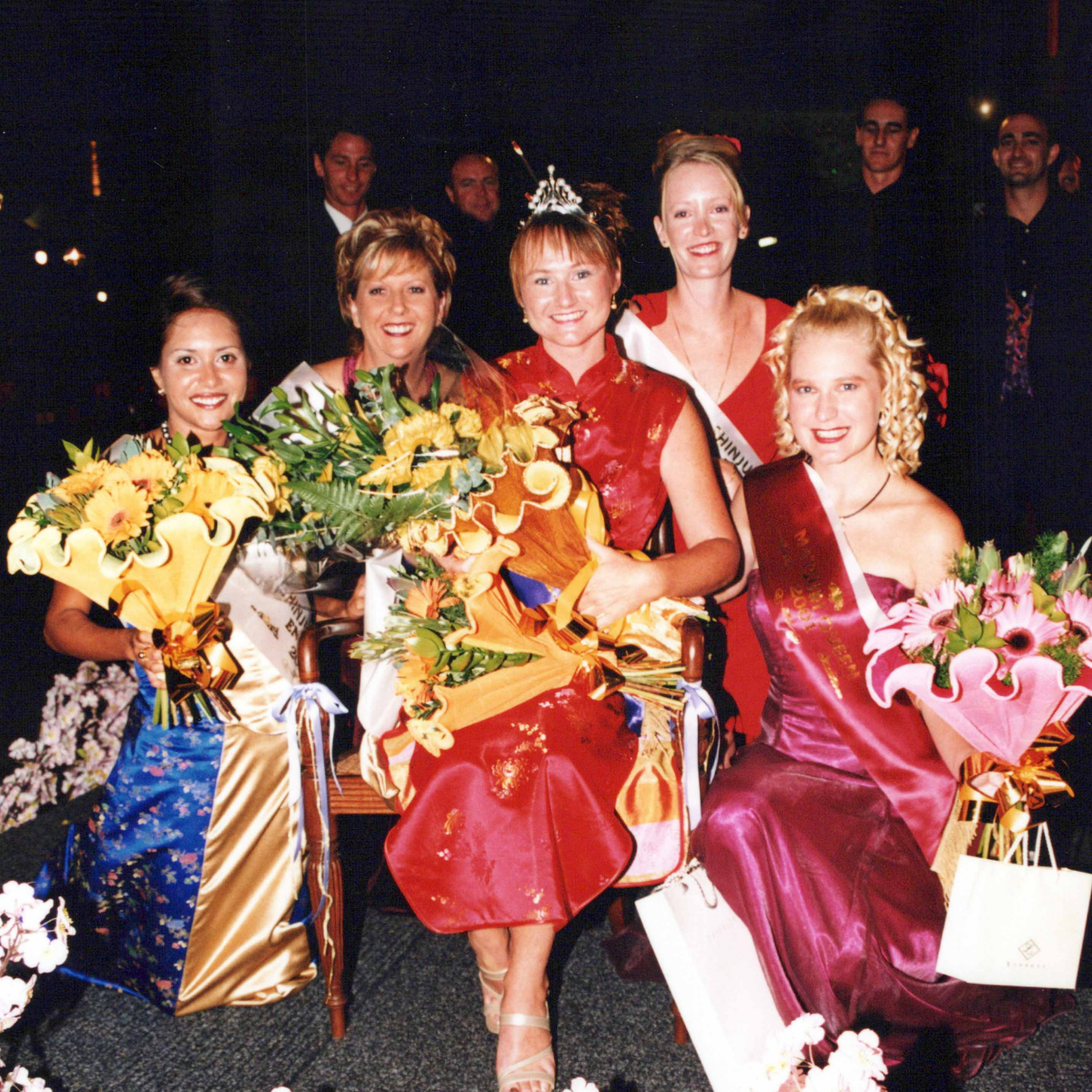 Five women posing together in formal evening wear for Shinju Matsuri celebrations in Broome, holding large flower bouquets. The central seated woman wears a red traditional-style dress and a tiara, while the others wear satin evening gowns in blue, red, and magenta.
