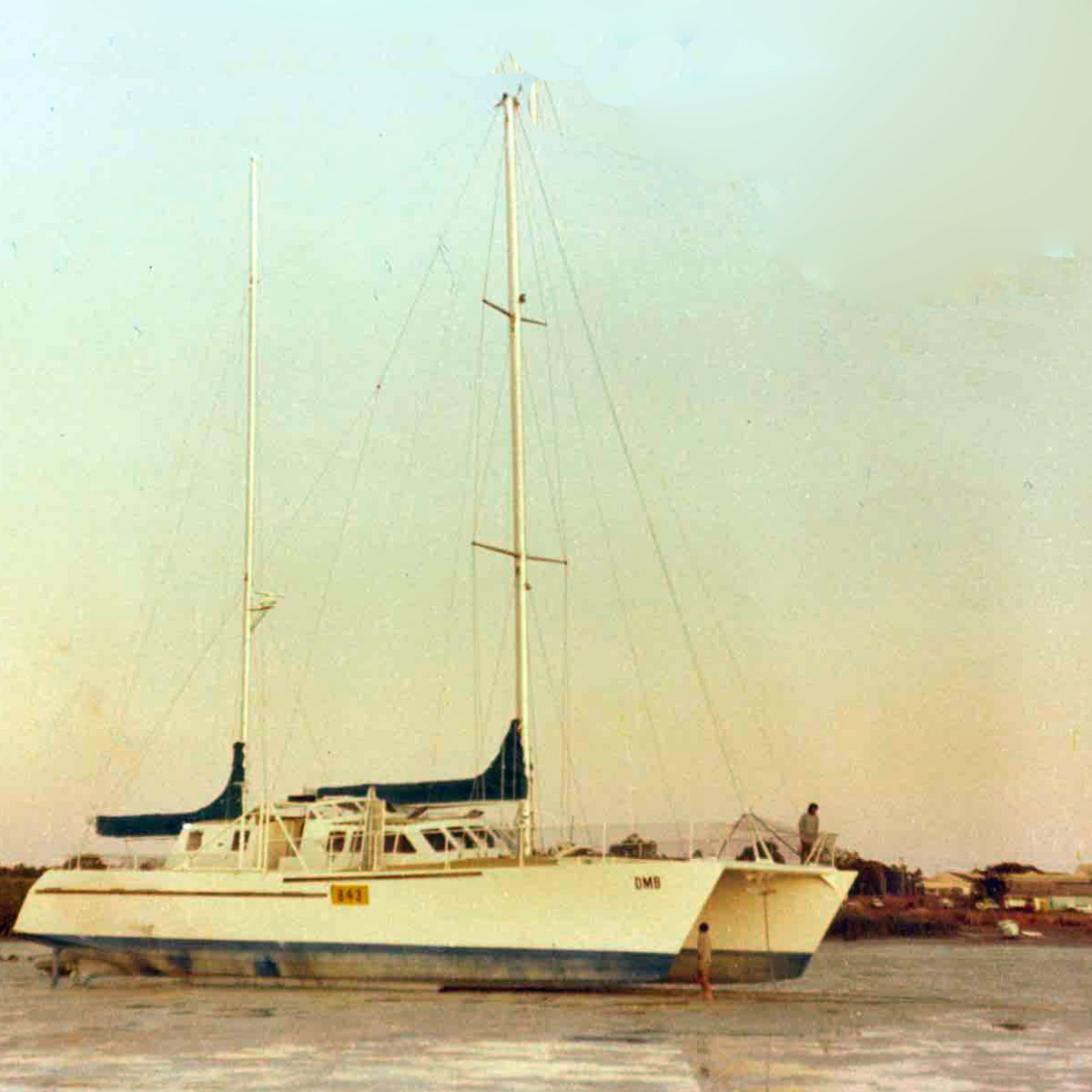 A white catamaran sailboat called the DMB with a blue hull stripe, photographed in the shallow Cygnet Bay water with its masts raised against a pale sky.