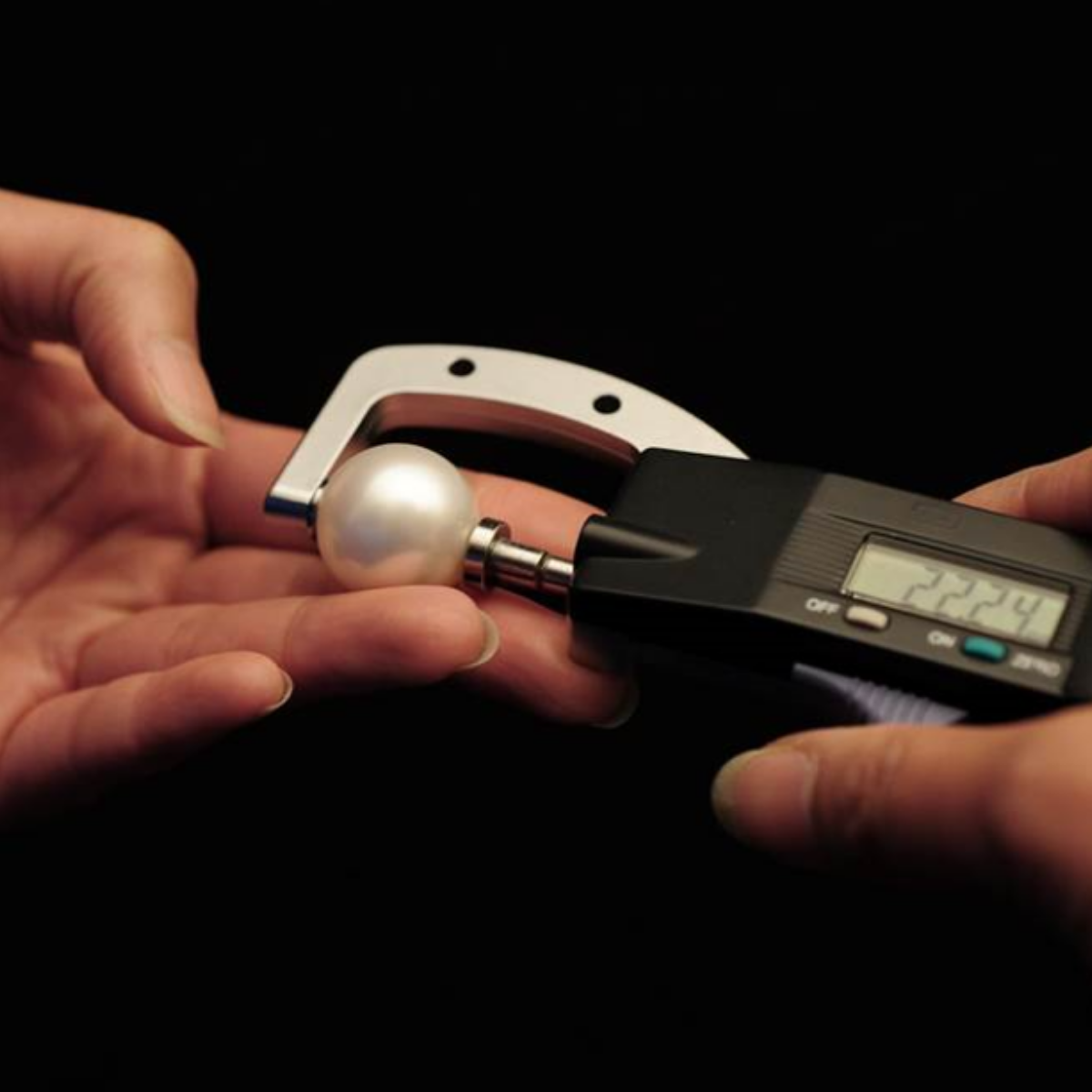 A close-up of a hand holding a large Australian South Sea white pearl under a digital micrometer caliper, with the display showing a measurement of 22.24 mm, against a solid black background. The largest fine quality, Saltwater South Sea pearl in history.